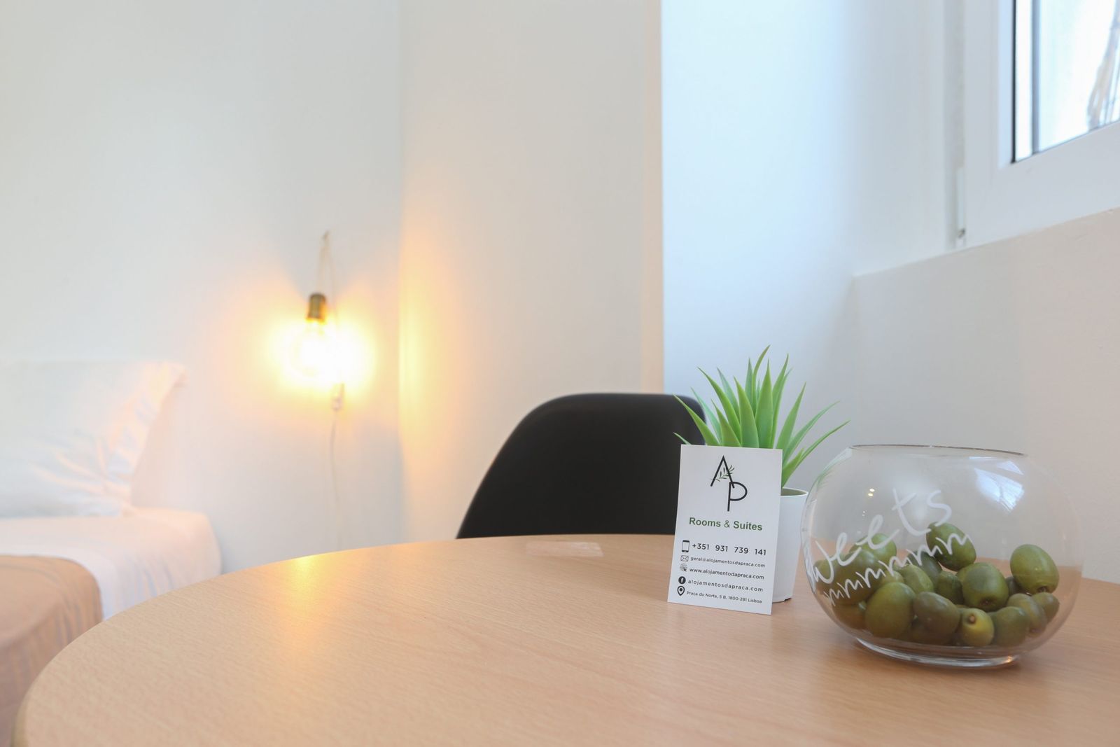 Quarto 5: mesa redonda com planta e taça de rebuçados, cadeira e candeeiro aceso junto à cama